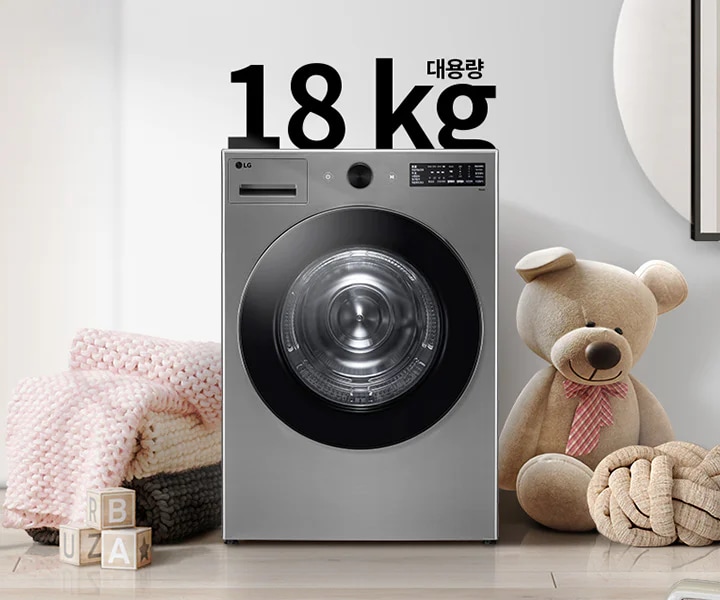 세탁기가 놓여 있으며, 세탁기 위에 대용량 18 kg이라는 텍스트가 있습니다. 세탁기 옆에는 곰 인형과 두꺼운 니트 담요가 놓여 있고, 바닥에는 나무 블록들이 있습니다. 배경은 밝고 현대적인 인테리어로 꾸며져 있습니다.
